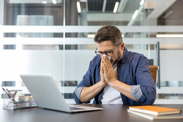 Obraz premium Businessman feeling unwell with cold or flu symptoms, blowing nose into a tissue while sitting at his office desk with a laptop, managing illness in a workplace environment