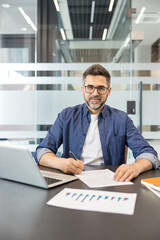 Obraz premium Professional businessman in smart casual attire at a modern office desk, smiling confidently while reviewing and signing documents with a pen beside his laptop and charts on screen