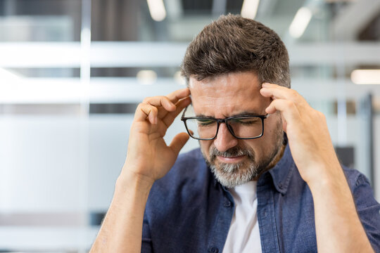 Mature man wearing glasses feeling intense pain, rubbing his temples while experiencing a severe headache or migraine, struggling with stress, burnout, and pressure in an office environment