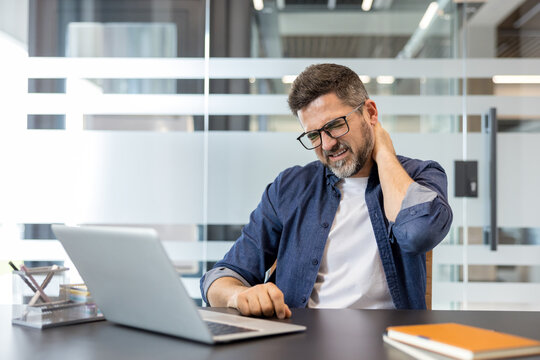 Man feeling intense neck pain and discomfort while sitting at his office desk, working on a laptop, indicating stress, poor ergonomics, and potential health issues from prolonged work