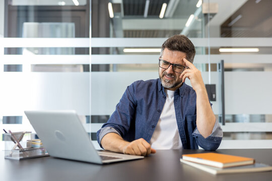 Mature man experiencing a severe headache and frustration while working diligently at his laptop, feeling overwhelmed and tired in a modern office environment