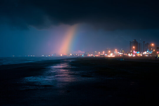 Glowing rainbow appearing over a sandy beach coastline and coastal town lights at night during a storm
