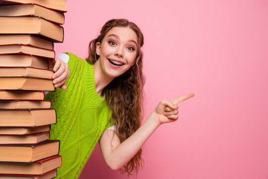 Young female student with braces smiles at pink background while peeking from a tall stack of book in a bright green sweater