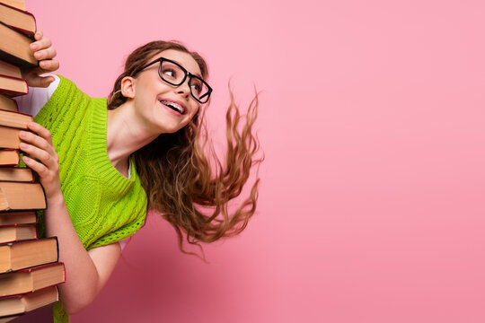 Young woman with glasses carries a stack of book smiling against a pink background