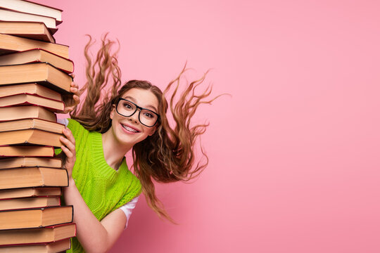 Young female student with glasses and a green vest peeks from a tall stack of book against a pink background smiling brightly
