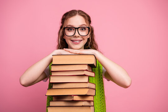 Happy young female student with glasses rests on a stack of book against a pink background smiling and showing study style