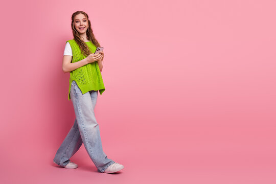 Young female student walks on pink background wearing bright green vest and jeans while using a phone