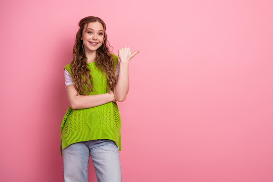 Young female student in a bright green knit vest smiles and points while standing against pink background
