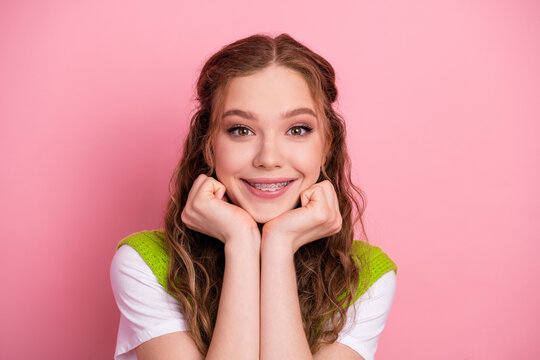 A young girl smiles with braces in a pink studio wearing a white shirt and a bright green vest