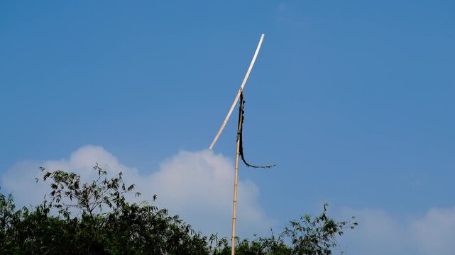 Traditional wooden propeller windmill on a bamboo pole rotating against a clear blue sky with white clouds and green trees in a rural setting.