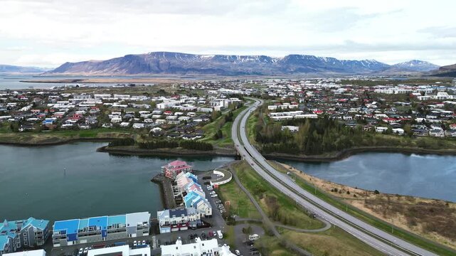 Drone panorama over Reykjavik waterfront suburbs with a lake, curving roads, colorful buildings and Mount Esja forming a dramatic background beneath clouds 4K.