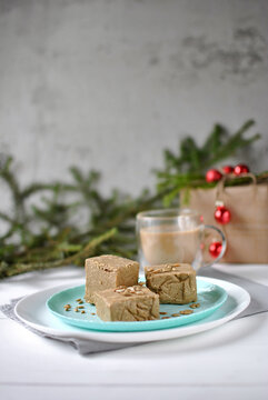 Sunflower halva cubes on a turquoise plate with tea, evergreen branches, red ornaments and a wrapped gift in a festive setting.
