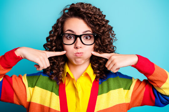 Playful young woman with glasses and curly hair wearing a colorful striped sweater making a funny face and pointing at her cheeks against a bright blue background