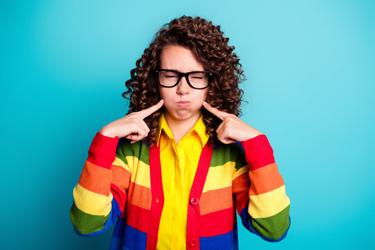 Young woman in rainbow cardigan pressing her cheeks and pouting with glasses, playful frustrated expression and colorful retro style against a turquoise background