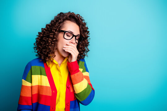 Tired woman yawning with glasses and curly hair wearing a rainbow sweater against a blue background