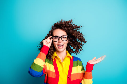 Happy young woman with curly hair and glasses wearing a colorful rainbow sweater on a bright blue background, smiling in a studio portrait while gesturing with palm up