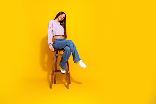 Smiling young woman sitting on a wooden stool against a bright yellow background wearing cropped sweatshirt and jeans &mdash; casual fashion portrait conveying relaxed, cheerful studio lifestyle