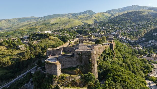 Aerial view of Gjirokaster Fortress with its iconic clock tower and historic stone walls surrounded by lush green hills and the city of Gjirokaster, Gjirokaster County, Albania.
