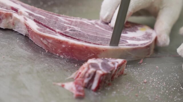 Close-up of knife cutting bone-in steak on stainless steel butcher table with gloved hands steadying meat, with meat, bone, knife, butcher detail, showing fat, marrow, steel, table context