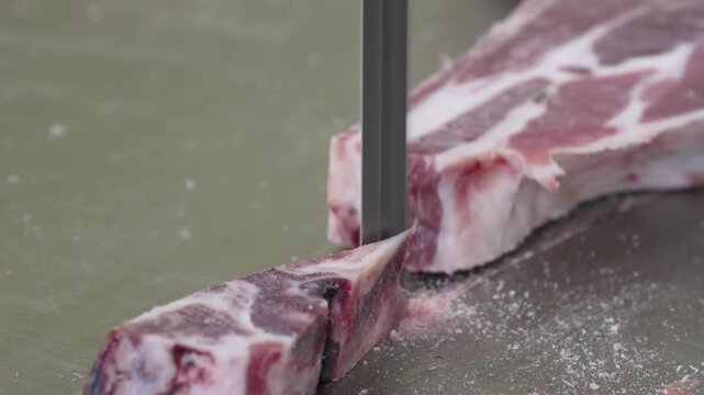 Gloved hand guiding vertical saw blade through thick raw pork rib near bone on metal work surface, with meat, pork, rib, blade detail, showing saw, butcher, cutting, slice context