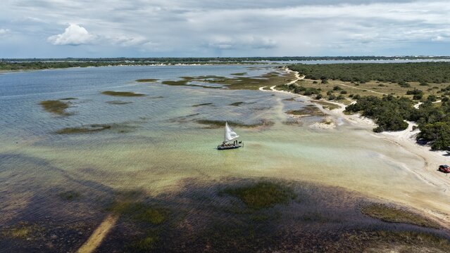Aerial view of a sailboat in the shallow turquoise waters of Parque Nacional dos Lencois Maranhenses, surrounded by coastal vegetation and sandy banks in Atins, Maranhao, Brazil.