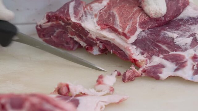 Gloved hands trimming raw pork with boning knife on cutting board, closeup diagonal blade slicing fat and connective tissue, with meat, pork, knife, glove detail