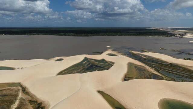 Aerial view of Parque Nacional dos Lencois Maranhenses featuring vast white sand dunes, turquoise lagoons, and lush vegetation along the river in Atins, Maranhao, Brazil.