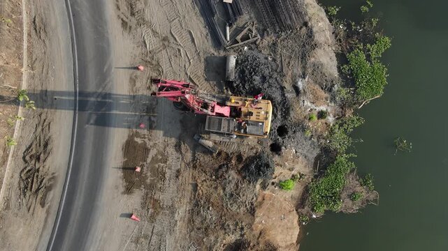 Aerial view of a construction site featuring a red piling machine on a dirt riverbank with a nearby road and motorcycle in Banyumas, Central Java, Indonesia.