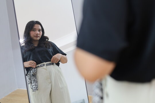 A young woman stands in front of the mirror at home, adjusting her clothes and trousers, getting ready for work.