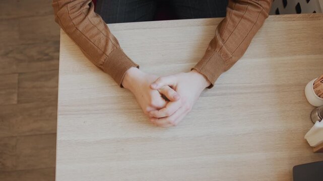 Close up of person hands resting on table occasionally interlacing fingers. scene includes wooden tabletop with condiment holders and napkin dispenser in soft natural light. Background