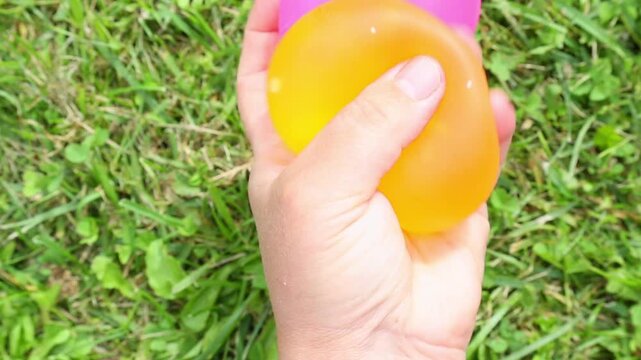 ASMR close-up of two hands pressing and squeezing pink and orange water balloons on green grass, gently reshaping the squishy textures in a slow satisfying relaxing motion.
