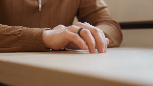 Close up of man hands resting on light wood surface fingers slightly moving with black ring visible. Man wears brown zip up sweater in soft indoor lighting background blurred to emphasize