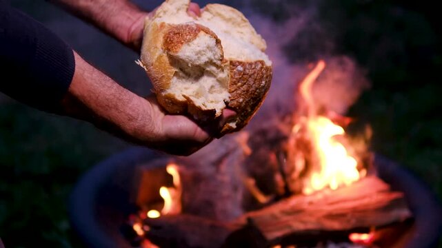 Male hands break apart a rustic loaf of white bread beside glowing flames and rising smoke at night, creating an authentic homemade bakery and artisan food atmosphere. Close up.