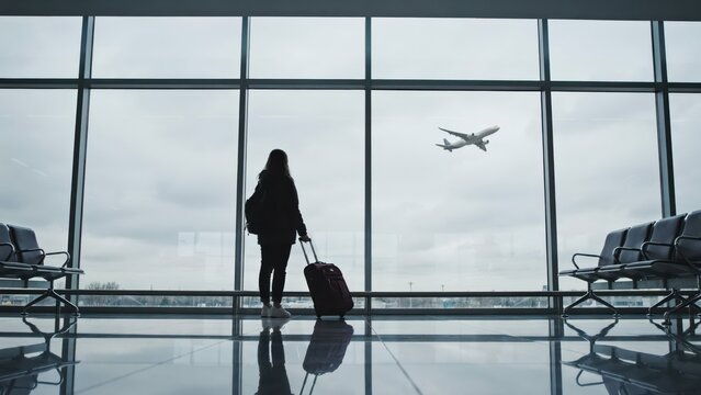 Silhouette of a woman traveler with luggage looking at an airplane from an airport terminal window.