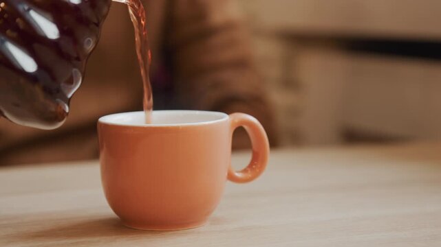 Close up of hand pouring dark liquid into empty peach ceramic cup on wooden table. action is steady and focused with background softly blurred to emphasize subject. Soft lighting highlights