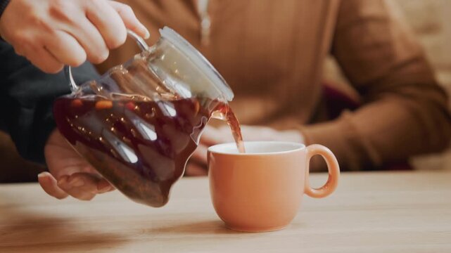 Close up of hand pouring dark liquid from glass carafe into peach colored cup on wooden table. person in brown clothing sits at background with hands resting on their lap throughout sequence