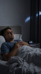 Fototapeta premium Caucasian man lies awake in bed at 3 AM looking troubled, suffering from insomnia. Mental health issue and sleep disorder concept.