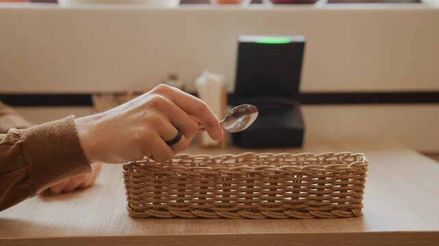 Close up of person placing silverware into woven wicker basket on wooden table. hand wearing brown long sleeve shirt and ring repeatedly places spoons from background towards empty