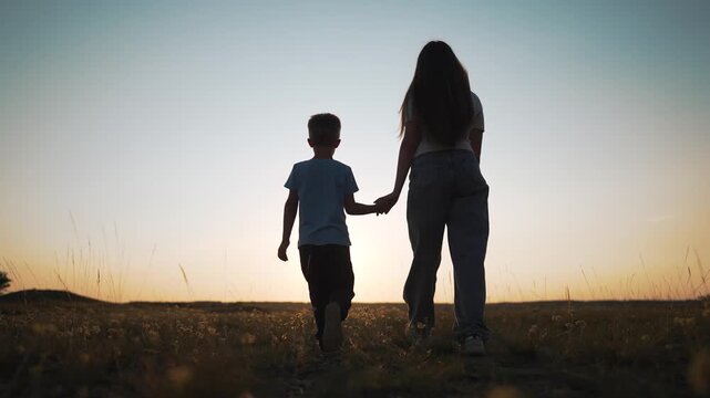 Mother and child walk holding hands in wildflower field at sunset. Boy walks with mother through field outdoors. Family in golden sunset light. Child and mother together walking in wildflower meadow.