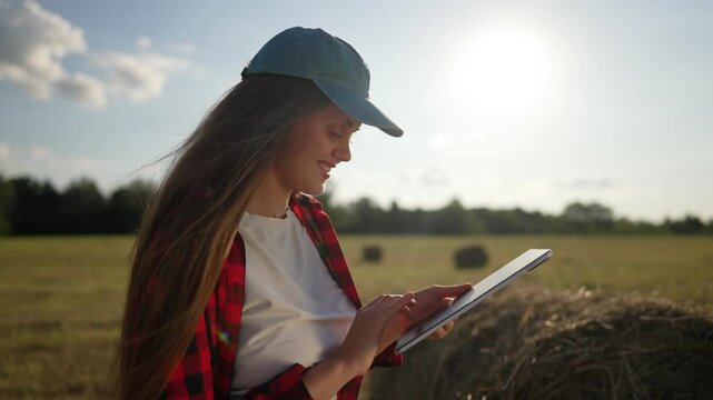 Smiling woman farmer uses tablet in sunny field near hay bale. Female worker smiles checking tablet data. Happy farmer works with tablet. Woman smiles using device in field. Farmer happy outdoors.