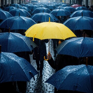 Bright yellow umbrella stands out among many blue umbrellas in rainy weather. Crowd of people walk on wet cobblestone street during heavy rain. Single yellow parasol individuality, uniqueness in sea