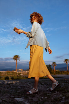 Casual woman walking outdoors holding phone, carefree mood, golden hour light, denim jacket, flowing skirt, relaxed lifestyle moment on a sunny day.