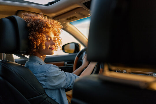 A woman driver sits behind the wheel in a sunlit car, denim shirt, curly hair framing her profile. A candid lifestyle portrait focused on independence and everyday travel.
