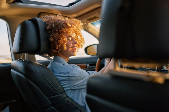 Woman with curly hair drives a car, smiles warmly, enjoying a sunny drive, casual lifestyle moment, interior shot illuminated by natural light.