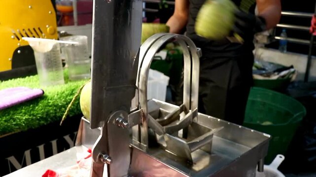 A street food vendor's gloved hand cuts a fresh young coconut using a stainless steel machine at a Thai night market, extracting pure coconut water for a healthy tropical beverage business.