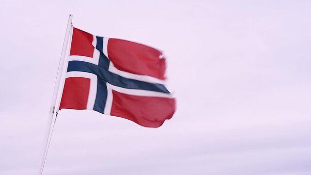 Norwegian flag snapping in wind against blank pale sky, Lofoten car ferry, Nordland, Norway