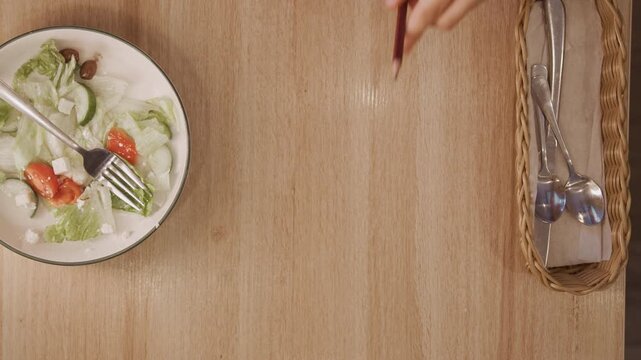Woman hands reach for salad bowl and utensils on wooden table placing them aside as woman positions empty notebook. scene features fresh vegetables in dish alongside neatly arranged