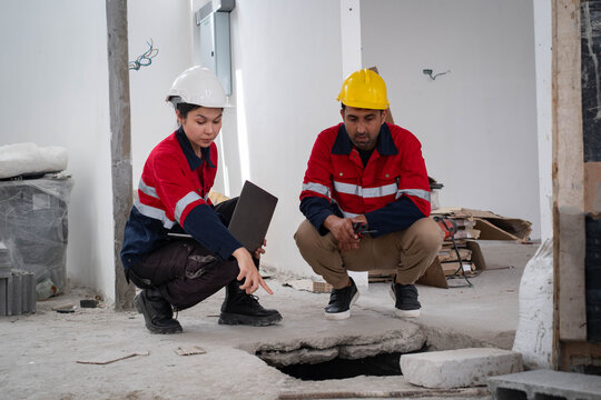Construction engineers inspecting a damaged concrete floor and foundation opening with a laptop during building assessment, highlighting structural inspection, workplace safety, renovation planning.