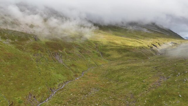 Aerial view of a lush green mountain valley with a small stream flowing through the landscape under low hanging clouds in Tromso, Troms, Norway.