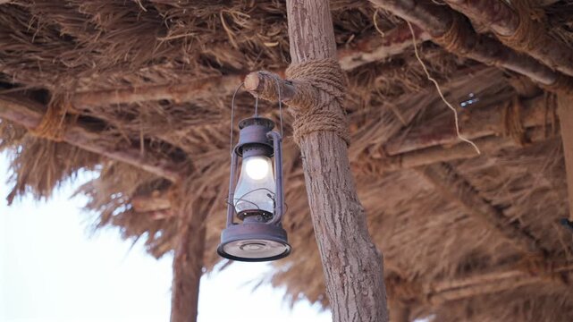 Close-up shot of glowing vintage kerosene lamp hanging from rustic wooden beam under thatched palm straw roof